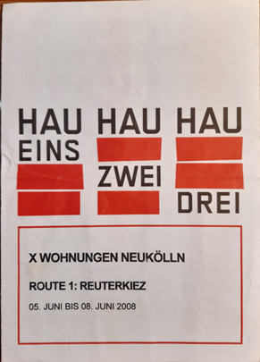 Auf dem abgebildeten Programmheft steht das Wort "HAU" drei mal nebeneinander und darunter in drei weiteren Zeilen jeweils das Wort "Eins", "Zwei" und "Drei". Außerdem sind die worte mit roten Quadraten angeordnet wie ein Schachbrett.